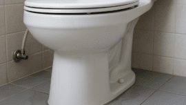 Close-up of a white ceramic toilet leaking water at the base onto a tiled bathroom floor.
