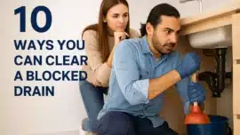 Plumber using a plunger under a kitchen sink while a concerned woman watches, illustrating a hands-on approach to clearing a blocked drain at home.